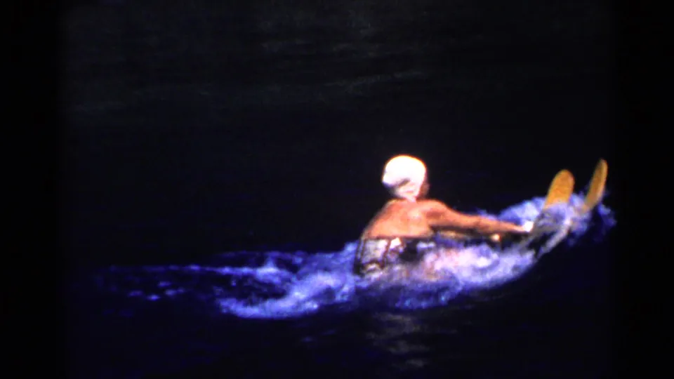 1955 California 8mm frame showing a woman in a white swim cap and dark swimsuit riding yellow water skis over dark water.