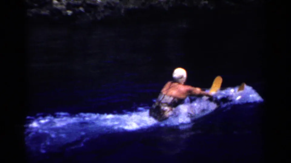 A woman water skier in a white cap leans forward as she is pulled across dark blue water in 1950s California home-movie footage.
