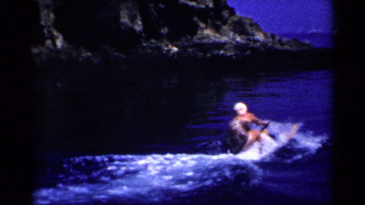 The water skier passes near a shadowed rocky shoreline, with blue water and bright spray visible in the 8mm frame.