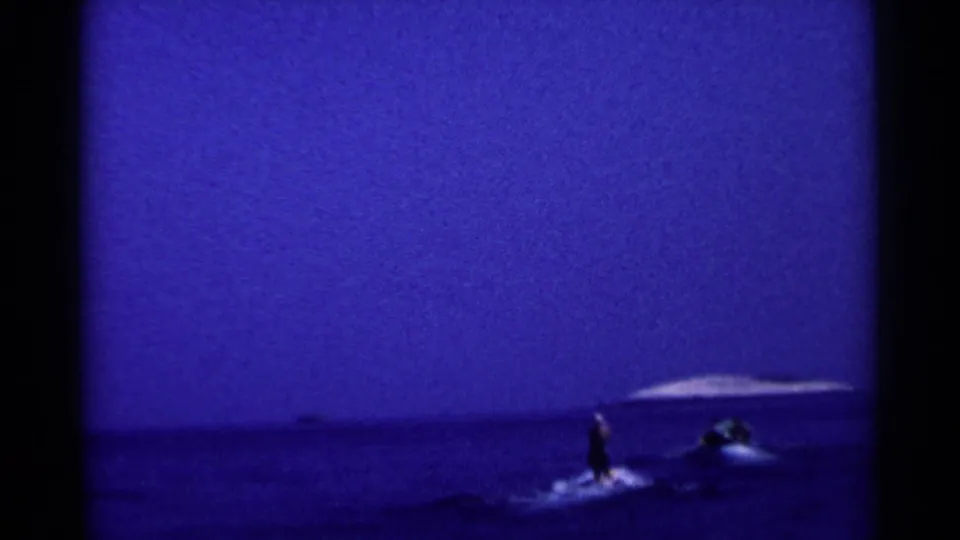 1955 California 8mm frame showing a distant water skier and small motorboat crossing dark lake water at dusk.