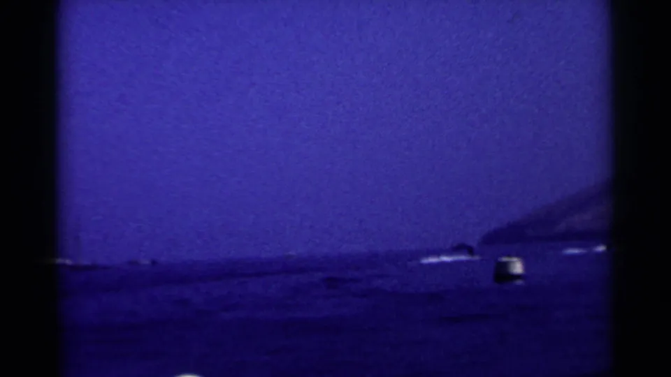 Grainy 1955 California lake view with a buoy in the foreground, distant boats, and hills along the shore.