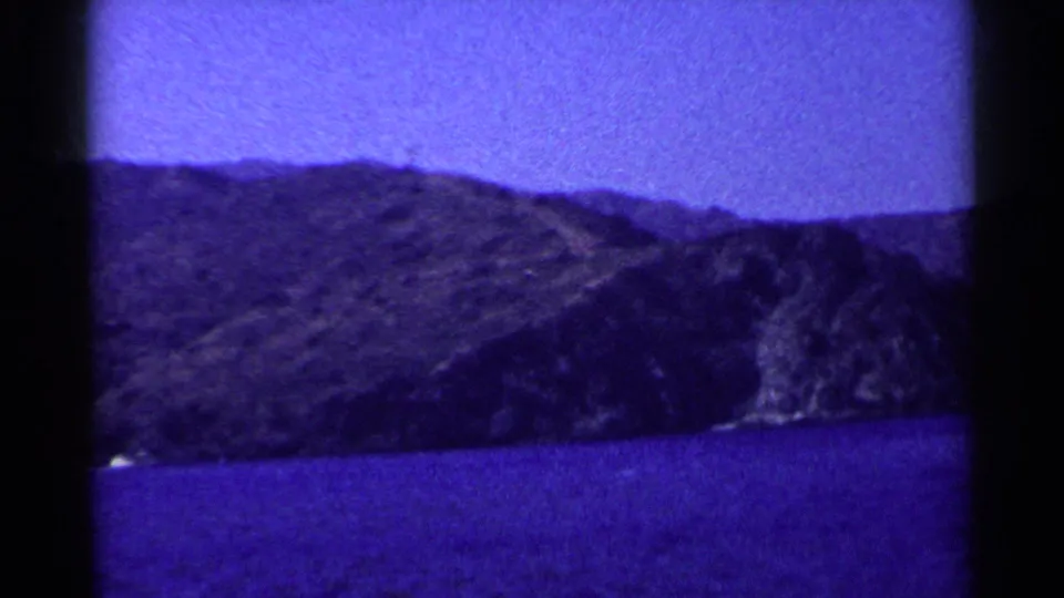 1955 California home-movie frame of blue water below rugged hills, with a small bright shoreline detail at lower left.