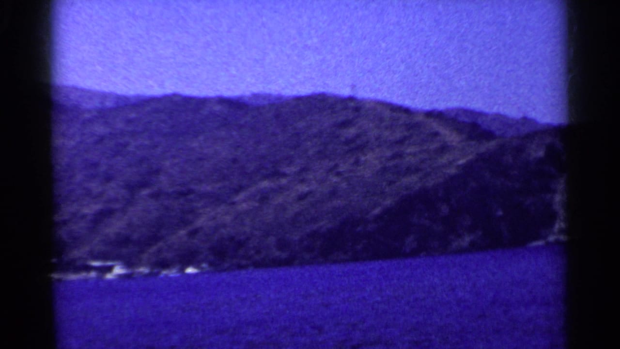 1955 California home-movie frame showing Lake Tahoe water, a wooded hillside, and distant shoreline structures under a purple-blue cast.
