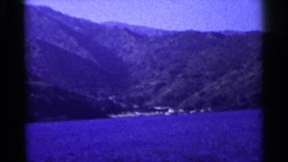 1955 California vacation frame showing Lake Tahoe water, wooded slopes, and faint shoreline structures in purple-blue 8mm color.
