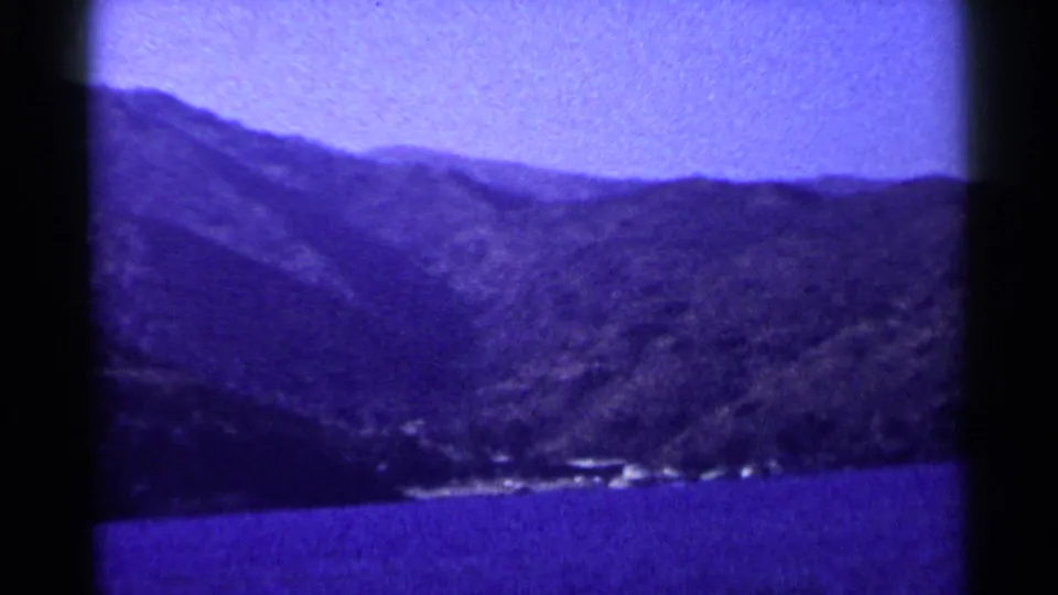 1955 California 8mm frame showing steep mountains behind a calm body of water with a small pale shoreline area.