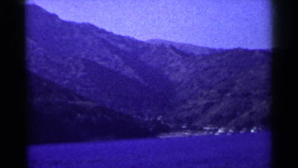 1955 California home-movie view of a mountainous shoreline seen across water, with soft focus and purple color cast.