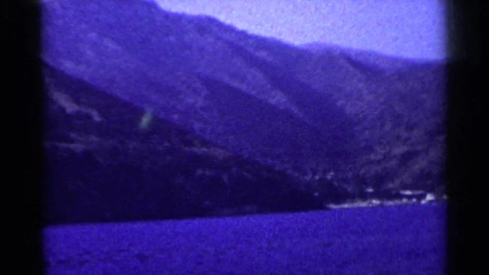 1955 California 8mm frame with layered mountain slopes, a narrow waterfront edge, and blue water in the foreground.