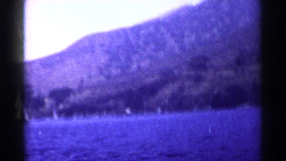 1955 California home-movie frame showing a bright sky, steep mountain slope, shoreline trees, and rippled water.