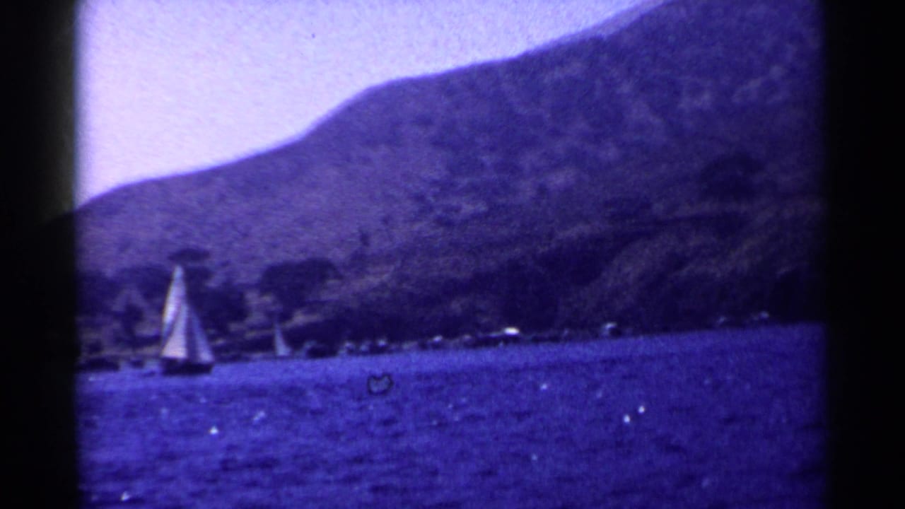 Small double-sailed boat on blue water with a mountain and shoreline behind it.