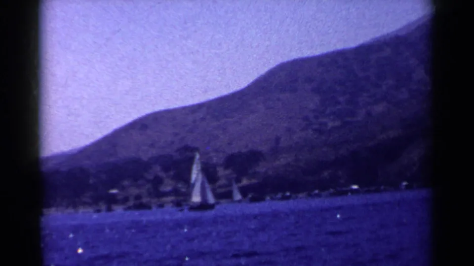 Vintage 8mm view of a double-sailed boat moving over blue water toward a mountain-backed California shore.