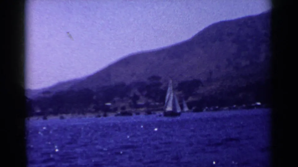 1955 California home-movie frame with a sailboat on sparkling water beneath a broad mountain slope.