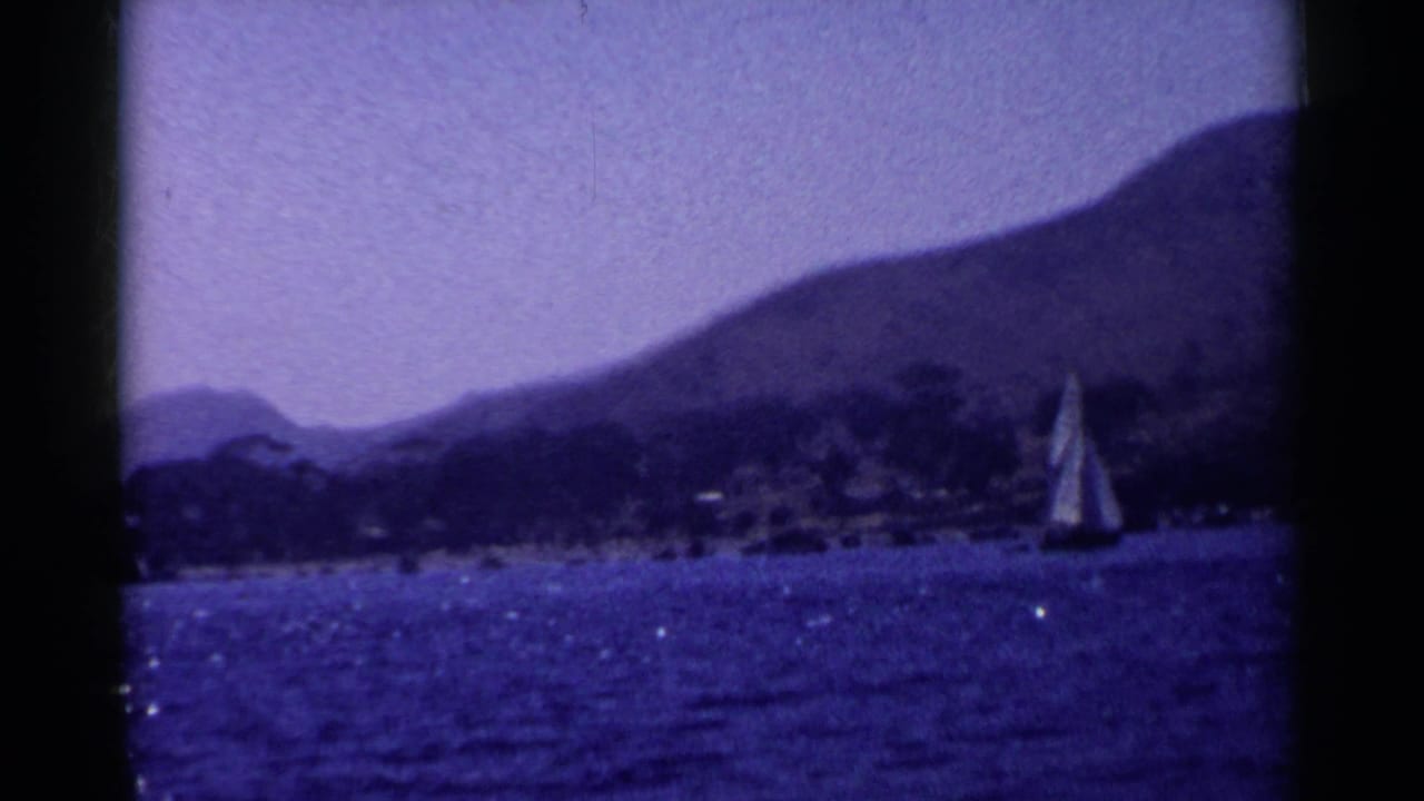 Sailboat on blue water with distant hills and shoreline trees.
