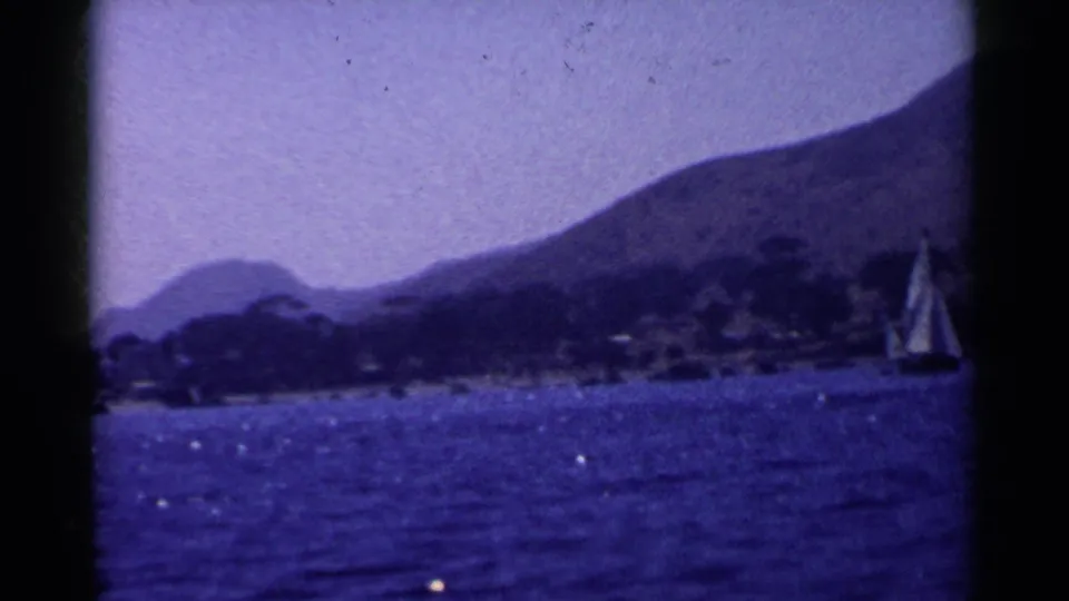 Vintage 8mm coastal view in California with a sailboat near the right side, blue water, shoreline trees, and hills.