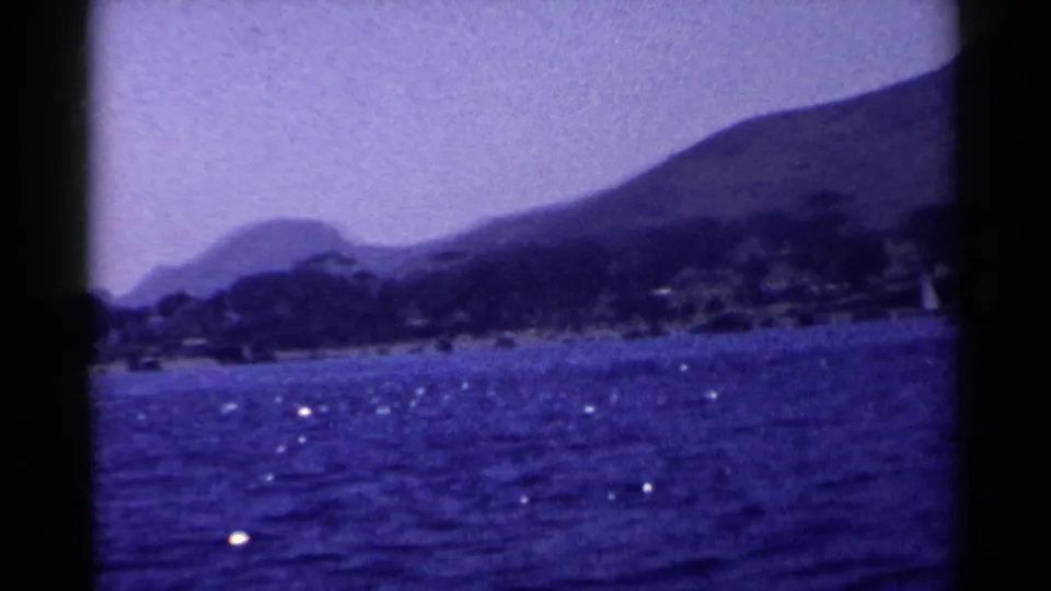 1955 California home-movie frame with sparkling ocean foreground, distant shoreline, hills, and a small sail partly visible.