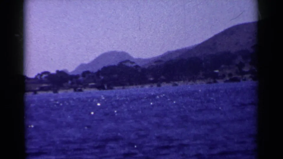 Vintage 8mm California coastal frame showing blue water, bright reflections, shoreline trees, and layered hills.