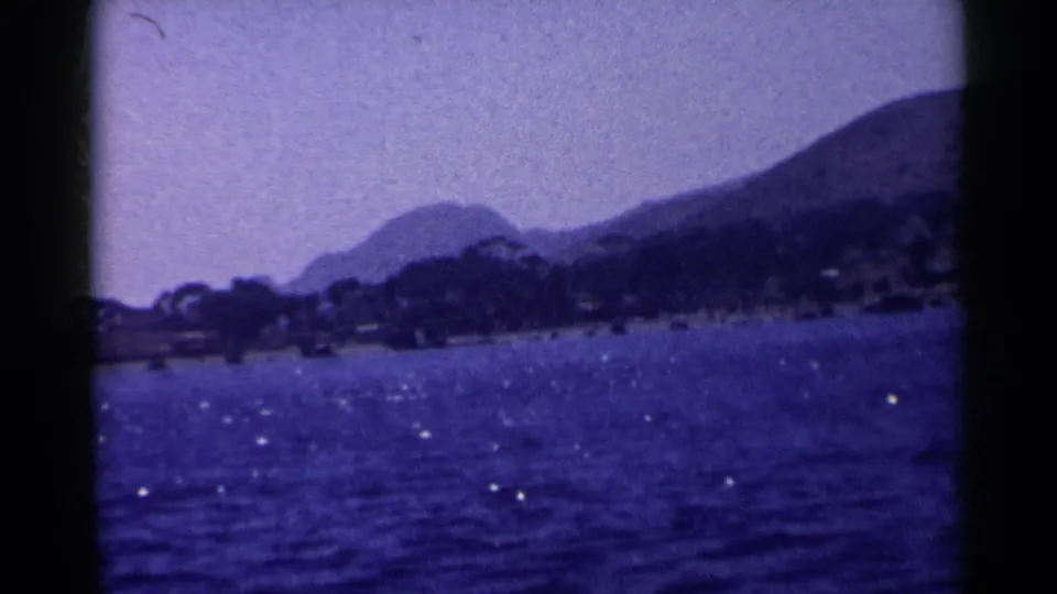 1955 California 8mm view across a blue lake toward a tree-lined shore and layered mountain silhouettes.