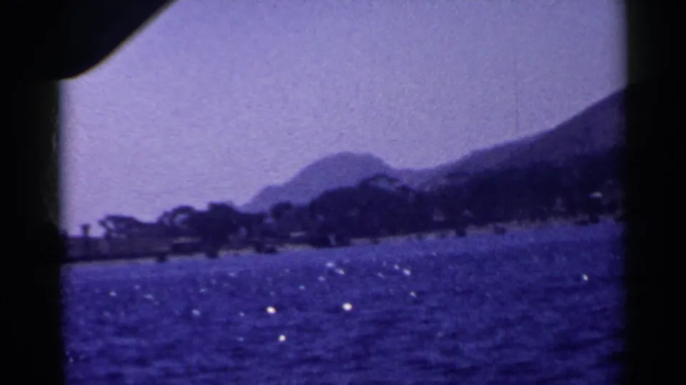 1955 California home-movie frame with blue lake water in front of a dark tree line and hazy mountain backdrop.