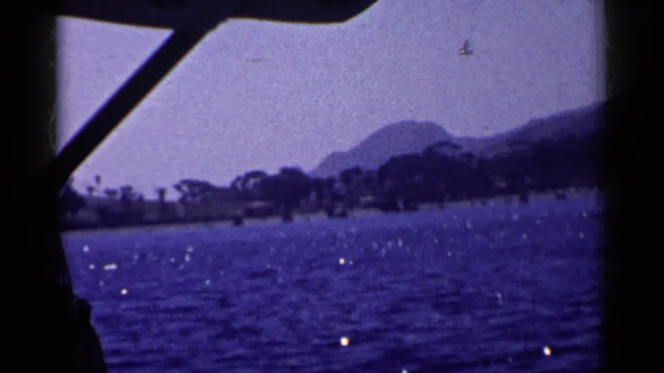 Boat structure silhouettes the 1955 California coastal view, with sparkling water, low shoreline, and hazy mountains beyond.