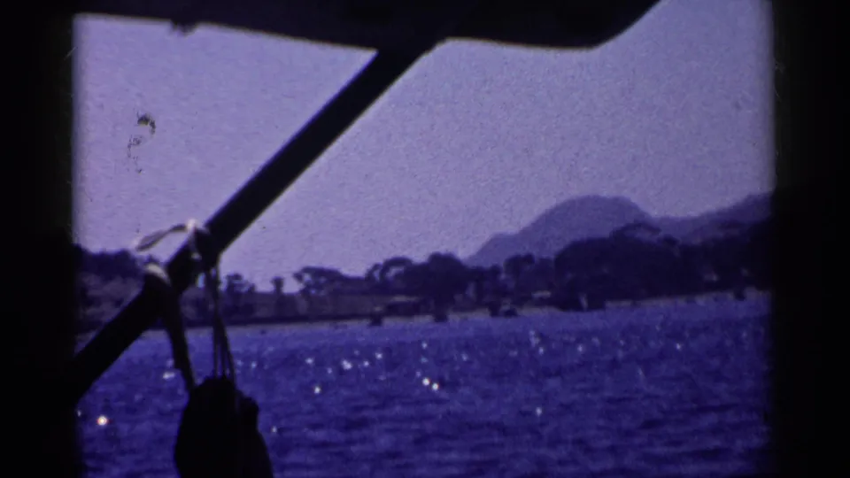 1955 California boating view with dark rigging in front of wavy water, a tree-lined shore, and a rounded hazy mountain.