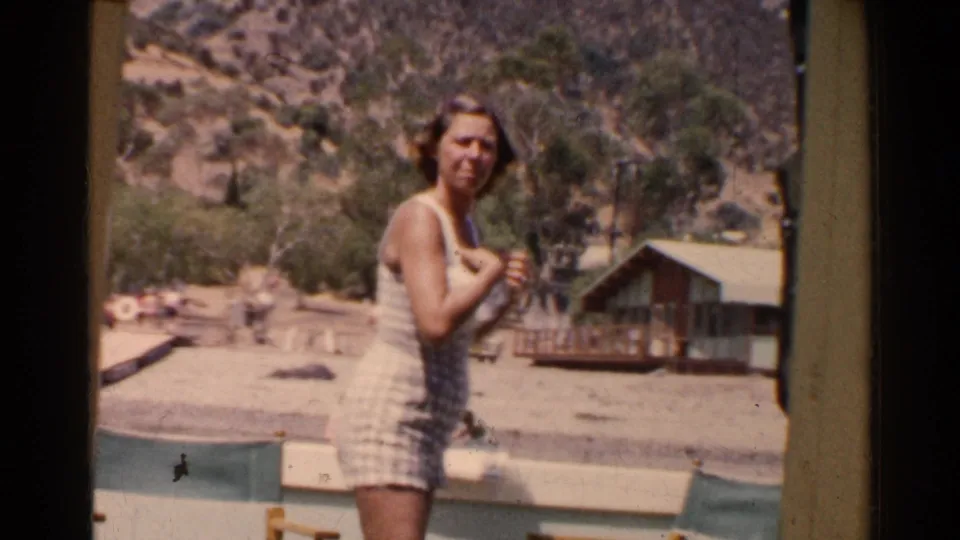 Woman in a white and blue checkered bathing suit stands at a California lakeside resort in 1955, with cabins and hills behind her.