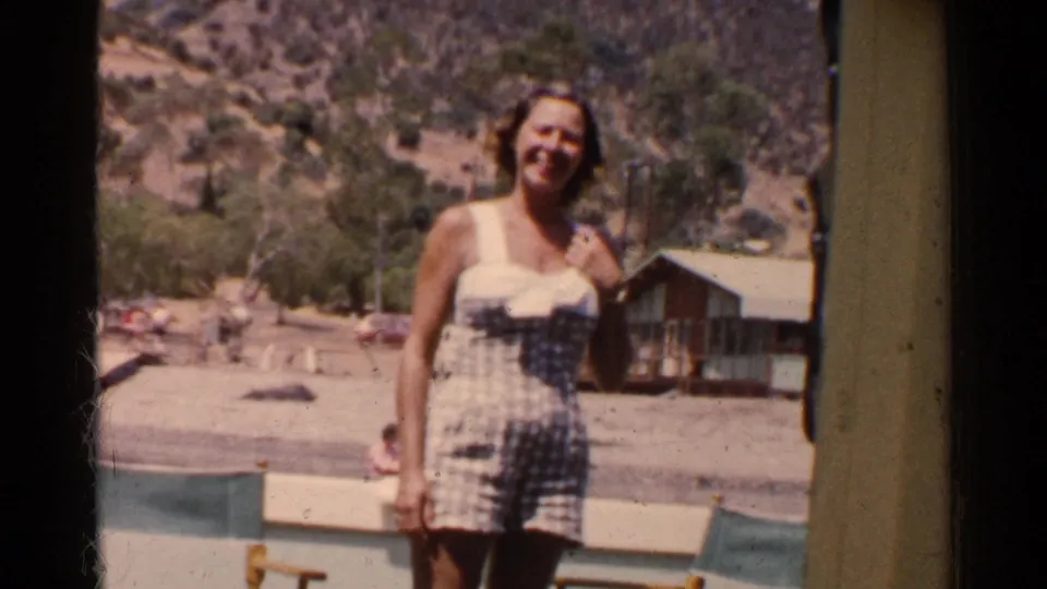 Smiling woman poses in a checkered bathing suit at a California resort, with a cabin, trees, and hillside visible behind her.