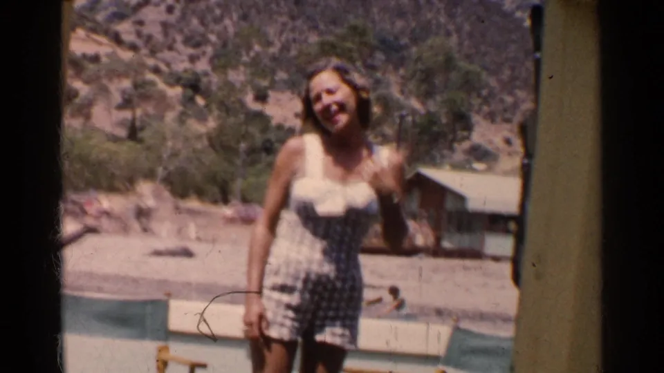 Woman smiles and raises a hand near a pale resort railing, with a cabin, trees, and California hillside in the background.