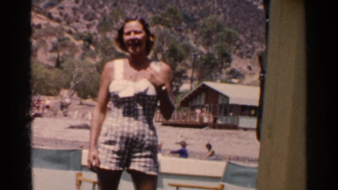 Young woman in a checkered 1950s swimsuit smiles near a California lakeside cabin with bathers in the background.