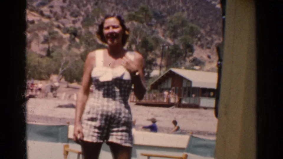 Young woman in a checkered 1950s swimsuit smiles near a California lakeside cabin with bathers in the background.