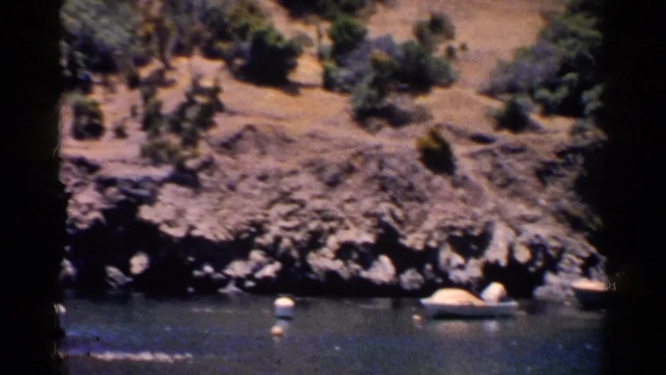 California lake shoreline with rocky banks, floating buoys or boats, dry hills, and scattered trees in a 1950s home-movie frame.