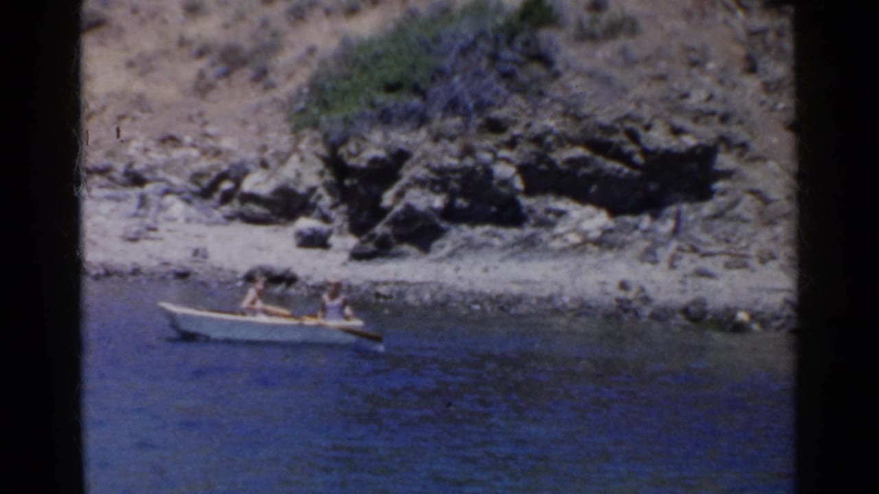 Two people in a small rowboat near a rocky lakeshore.