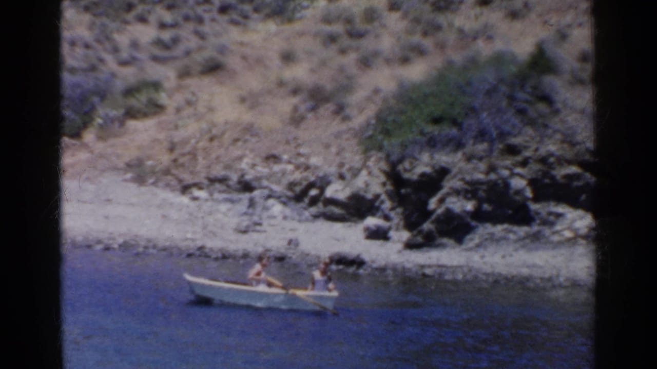 Small rowboat with two people moving along blue water near shore.