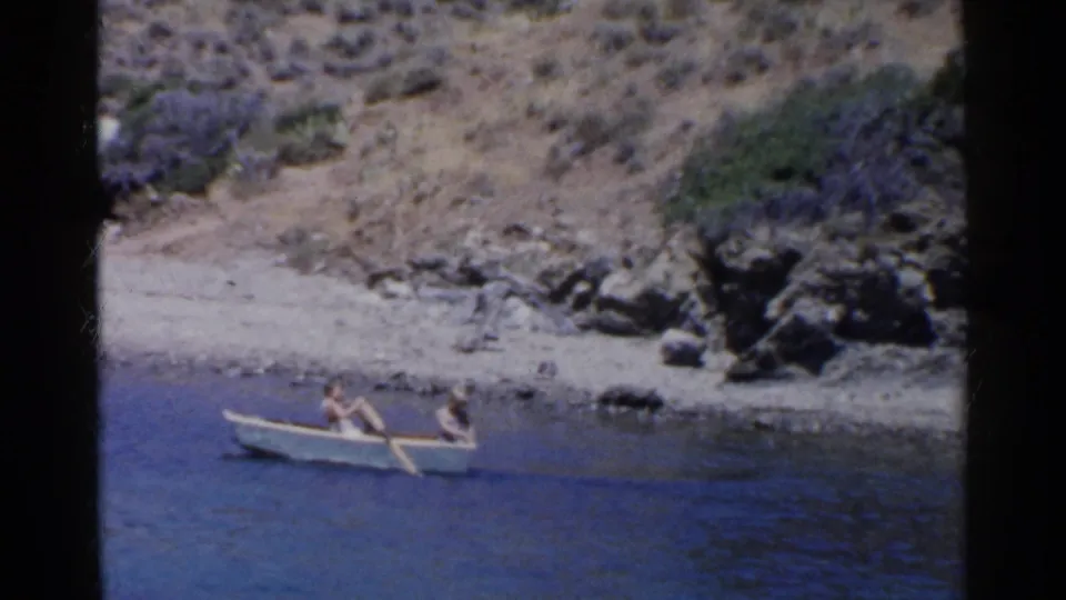 1955 California 8mm frame showing a small rowboat with two people near rocks, scrub, and blue water.