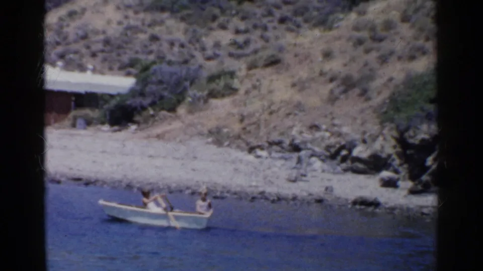 1955 California home-movie frame showing two people in a rowboat, with rocky shore and a small structure in the background.