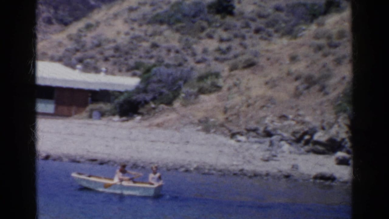 1955 California 8mm frame showing two people in a rowboat on blue water below a wooden shoreline house.