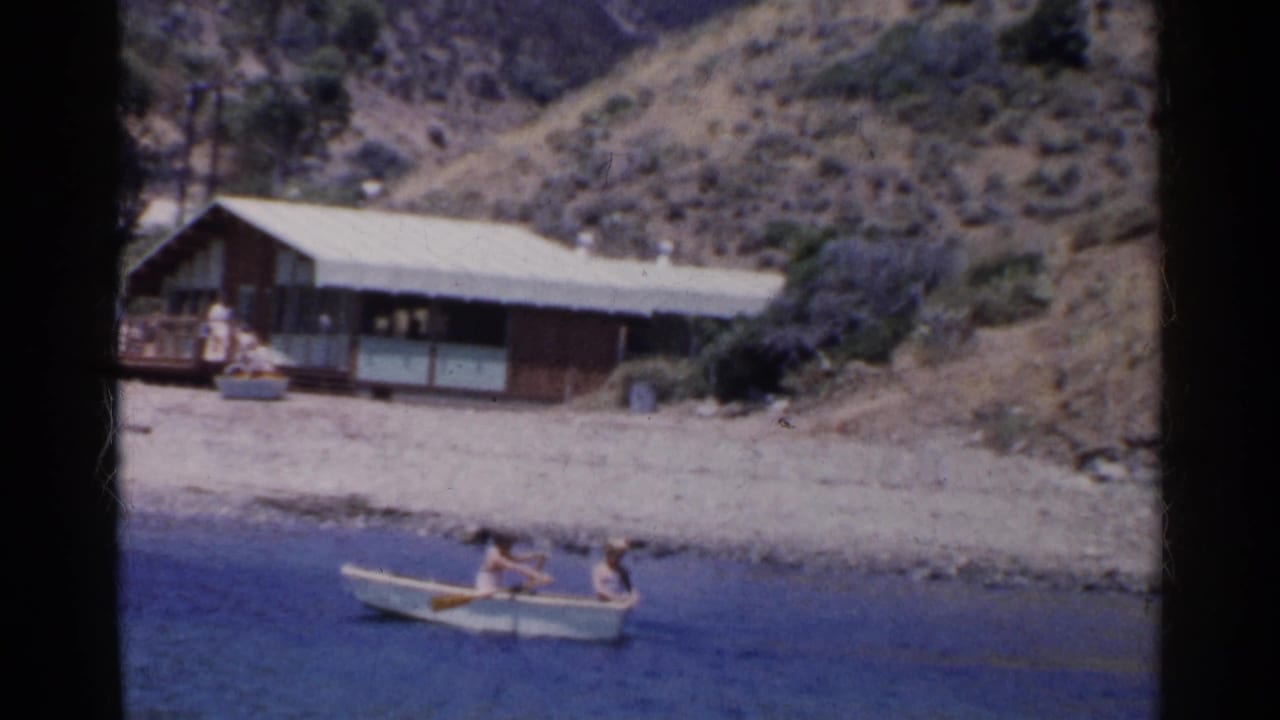 Two people row a small white boat along the shore in a 1955 California home-movie frame.