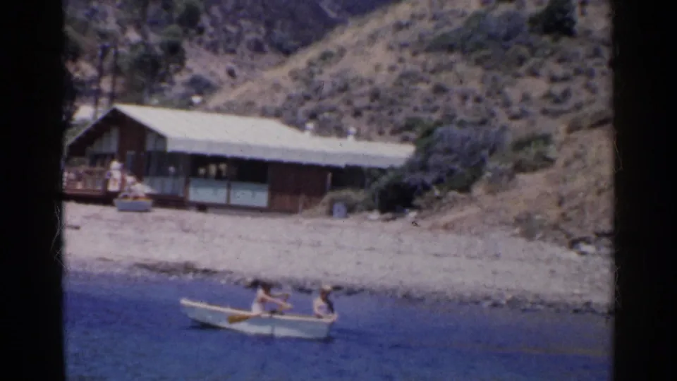 Two people row a small white boat along the shore in a 1955 California home-movie frame.