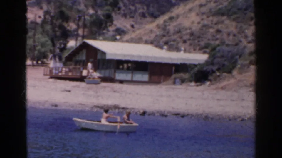 A 1955 California shoreline view shows two people rowing below a rustic house and dry hillside.