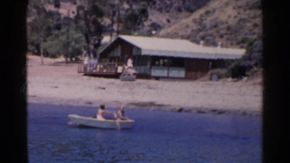 1955 California home-movie frame of two people rowing past a sandy shore with a wooden house behind them.