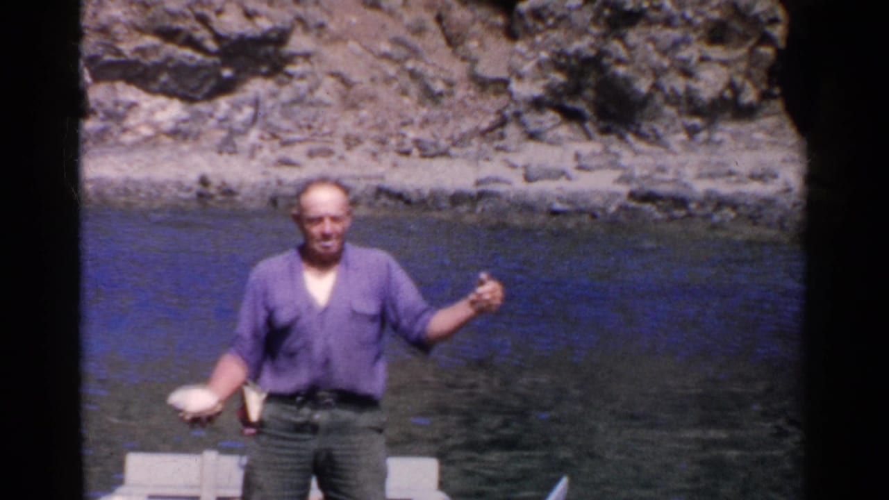 Man standing in a small boat on calm water near a rocky shoreline.
