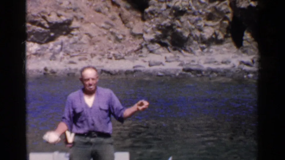 In this 1955 California home-movie frame, a man faces the camera from a small boat while holding a small white object near calm water.