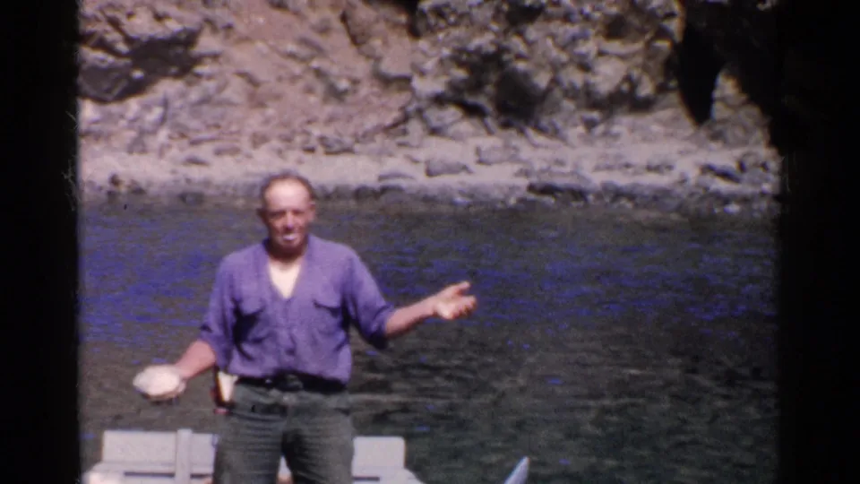A man in a purple-blue shirt gestures with both hands from a small boat beside calm water and a rocky California shoreline.