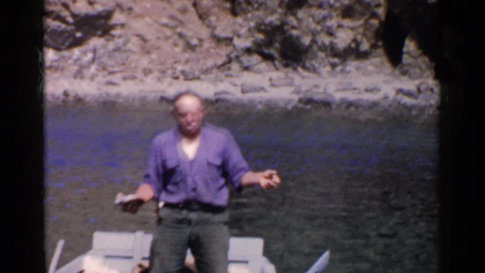 The man remains standing in the small boat, lowering one hand as calm water and a rocky California shoreline sit behind him.