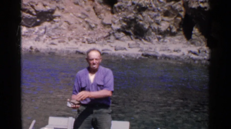 In 1955 California home-movie footage, a man in a purple shirt sits in a small boat near a rocky cliff while holding clams.