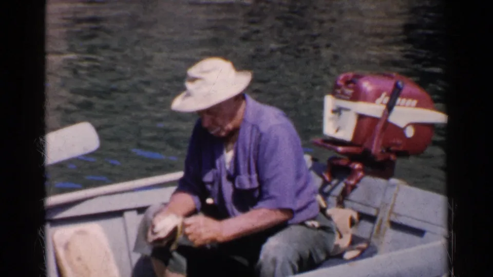 A man in a white hat and purple shirt cleans clams by hand in a small boat with a red Johnson outboard motor visible behind him.