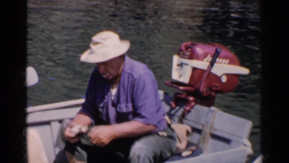 Close 8mm view of a man seated low in a small boat, cleaning clams while a red outboard motor and rippled water fill the background.