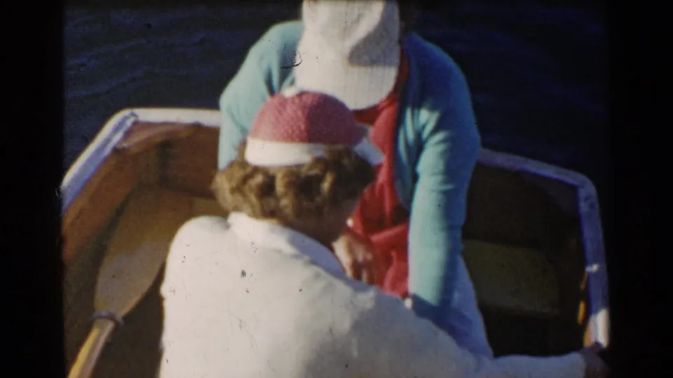 1955 California home-movie frame showing two women in a wooden rowboat, viewed from above, with lake water around them.