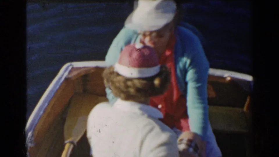 1955 California home-movie frame of two women seated in a wooden rowboat, with one woman in a white cap facing the camera area.
