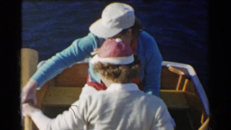 1955 California home-movie frame of a woman in a blue cardigan reaching across a wooden rowboat while another woman sits in front of her.