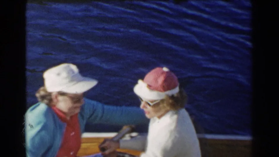 In metadata-supported 1955 California home-movie footage, two older women sit close together in a small wooden boat, one in a white cap and turquoise cardigan and the other in a red-and-white sun hat.