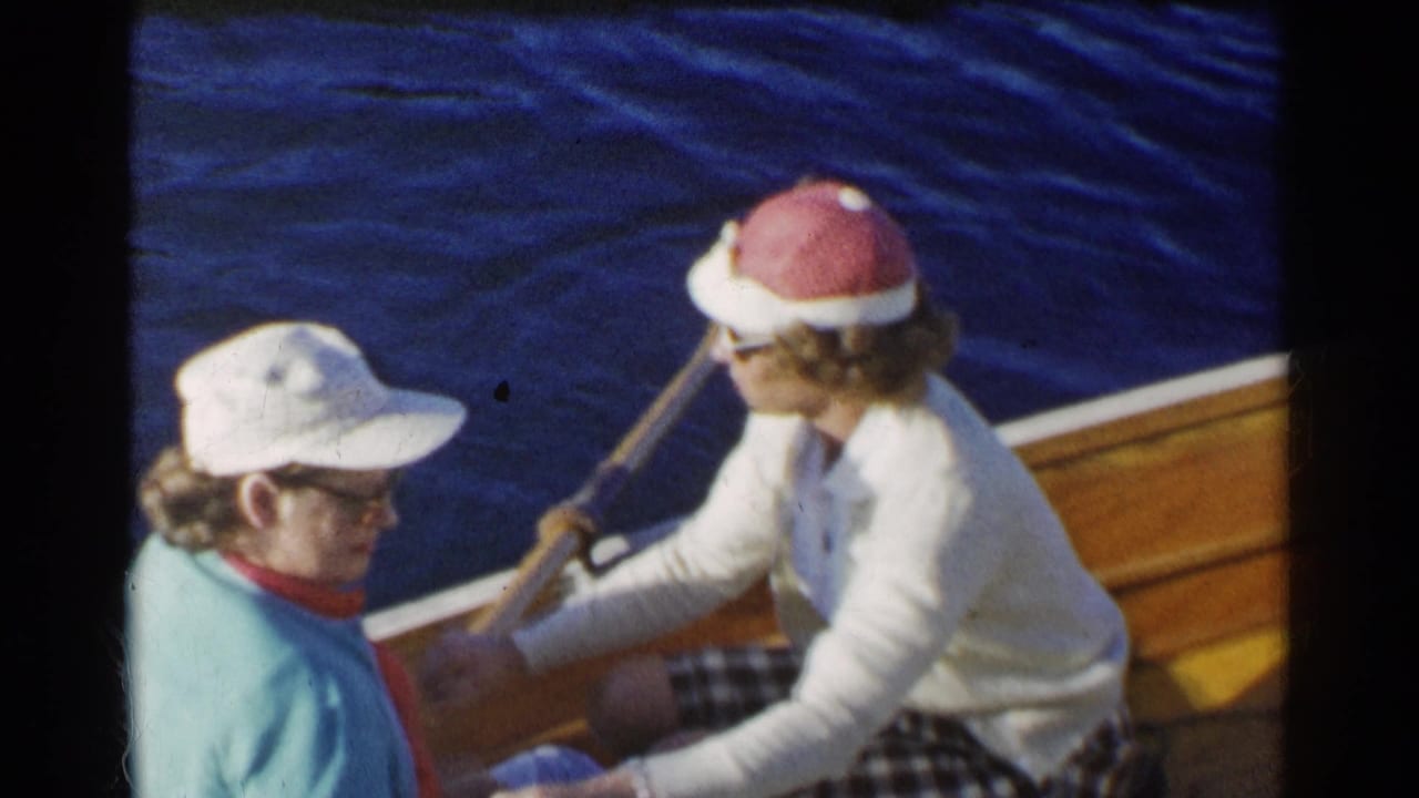 Two older women sit in a wooden boat as one woman holds a paddle near the water.